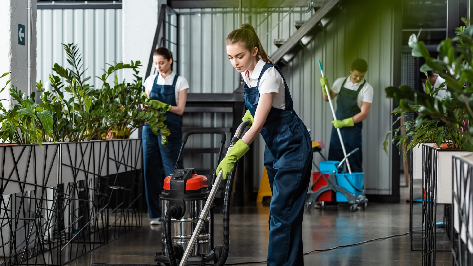 A group of three people in uniforms cleaning an indoor space filled with plants. One person is vacuuming with a wet/dry vacuum, while the others are tending to plants. The setting features a modern, well-lit environment with stairs in the background.
